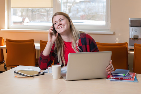 A smiling blonde woman sitting at a laptop and talking over a cell phone.の写真素材