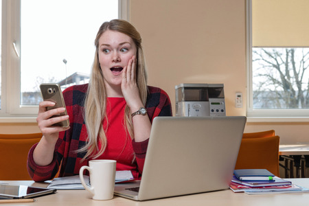 Smiling blonde woman, who is sitting at a laptop and surprised looks at the phone.の写真素材