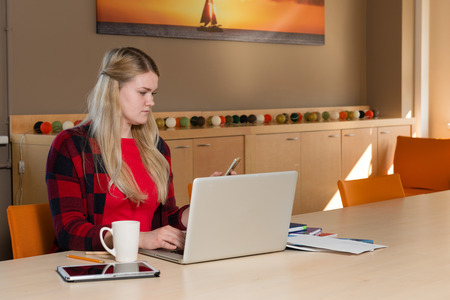 Smiling blonde woman sitting at a laptop drinking tea and working.の写真素材