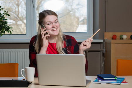 A blonde woman sitting at a laptop and talking over a cell phone.の写真素材