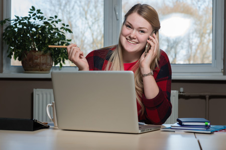 A smiling blonde woman sitting at a laptop and talking over a cell phone.の写真素材