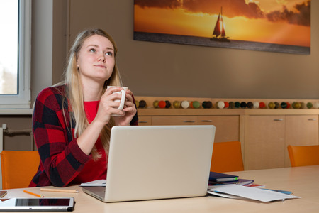 Smiling blonde woman sitting at a laptop drinking tea and working.の写真素材