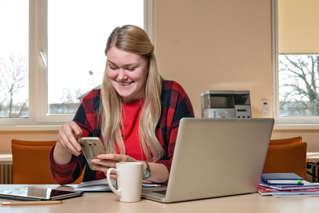 A smiling blonde woman sitting at a laptop and talking over a cell phone.の写真素材