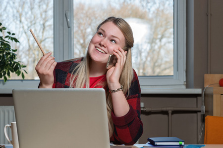 A smiling blonde woman sitting at a laptop and talking over a cell phone.の写真素材