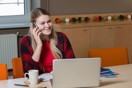 A blonde woman sitting at a laptop and talking over a cell phone.の写真素材