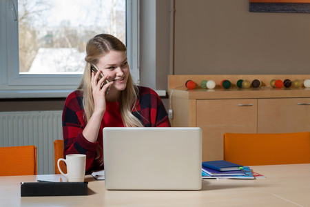 A blonde woman sitting at a laptop and talking over a cell phone.の写真素材