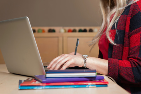 Blonde woman sitting at a laptop and writing. There is a laptop, a tablet, a phone and a notebook on the table.の写真素材