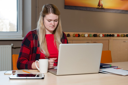 Smiling blonde woman sitting at a laptop drinking tea and working.の写真素材
