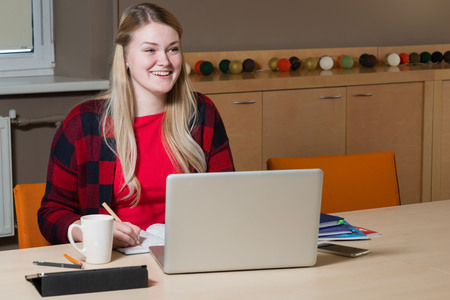 Smiling blonde woman sitting at a laptop drinking tea and working.の写真素材
