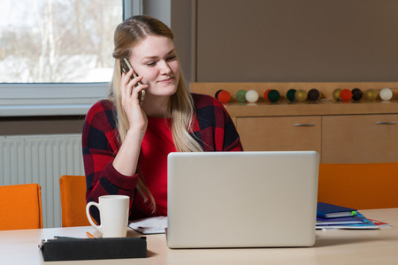 A blonde woman sitting at a laptop and talking over a cell phone.の写真素材