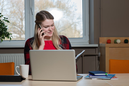 A blonde woman sitting at a laptop and talking over a cell phone.の写真素材