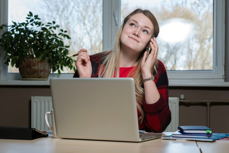 A smiling blonde woman sitting at a laptop and talking over a cell phone.の写真素材
