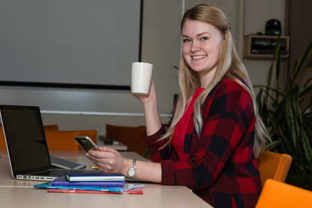 Smiling blonde woman sitting at a laptop drinking tea and working.の写真素材