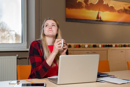 Smiling blonde woman sitting at a laptop drinking tea and working.の写真素材
