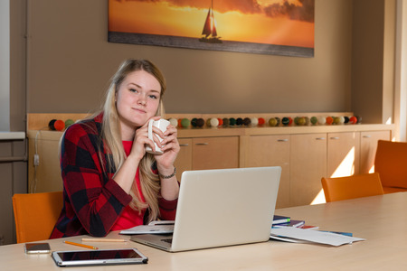 Smiling blonde woman sitting at a laptop drinking tea and working.の写真素材