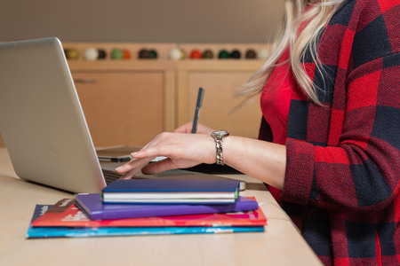 Blonde woman sitting at a laptop and writing. There is a laptop, a tablet, a phone and a notebook on the table.の写真素材