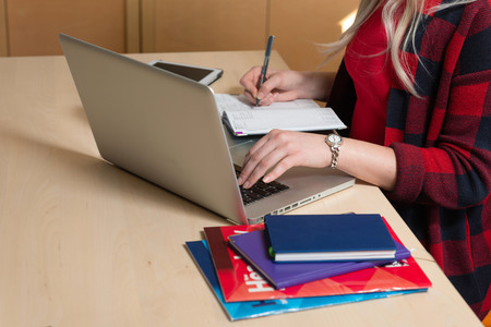 Blonde woman sitting at a laptop and writing. There is a laptop, a tablet, a phone and a notebook on the table.の写真素材