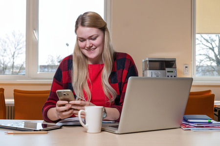 A smiling blonde woman sitting at a laptop and talking over a cell phone.の写真素材