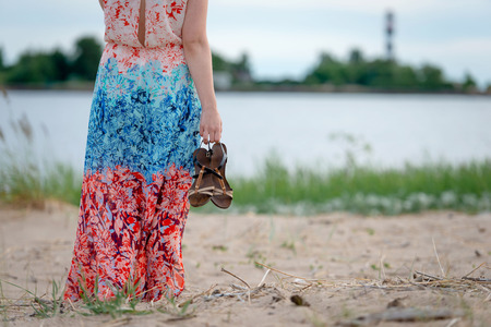A young woman wearing a colorful long dress and shoes in her hands standing on the beach and looking at the lighthouse. view from the backの写真素材