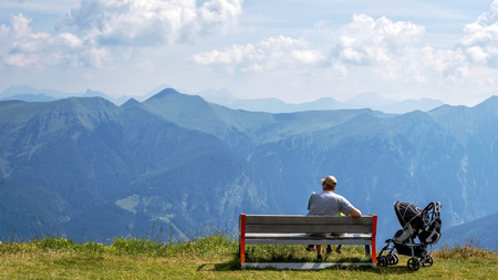 A man with a child sits on a wooden bench at the top of the Alps and looks at the beautiful mountain landscape.の写真素材