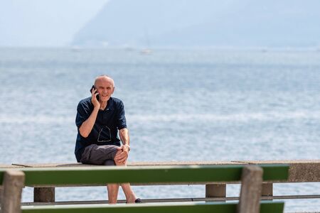 GMUNDEN, AUSTRIA, - AUGUST 03, 2018: The man sits at the edge of the lake promenade and talks over the phone.のeditorial素材
