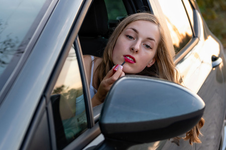 A young, beautiful woman with long hair through the car window looks in the rear view mirror and paints her lips.の写真素材