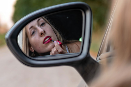 A young, beautiful woman with long hair looks in the car's rearview mirror and paints her lips.