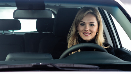 Blonde woman sits at a white car steering and seen through the window.