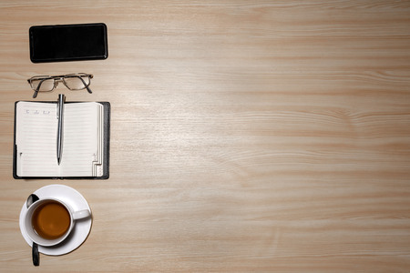 Desk with eyeglasses, notepad, smartphone, pen and a cup of tea on a wooden table. Top view with...