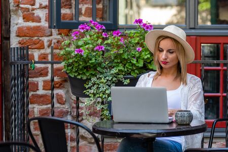 Beautiful blond woman with sun hat dressed in  light clothes sitting in outdoor cafe and working with computer.の写真素材