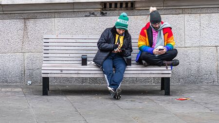 London, UK - January 1, 2020: Two men sit on a bench on the side of the street and look at their smartphonesのeditorial素材