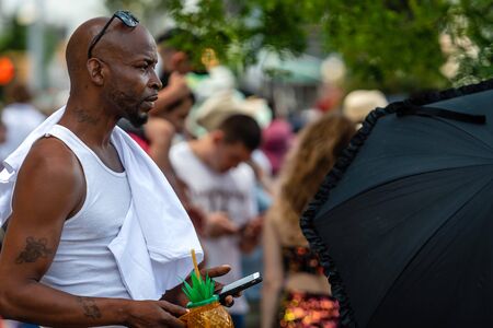 Brooklyn, New York City, USA - June 22, 2019: Coney Islands annual mermaid parade, african man with drink mug standing in crowd and looking at smartphoneのeditorial素材