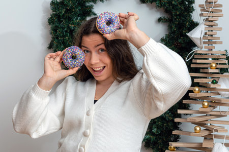 slim brunette in a white knitted jacket holding colorful donuts against her eyes, attractive young woman with long hair having fun with sweets, close-upの写真素材