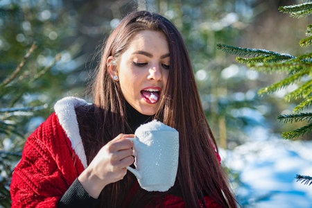 teenage woman with a red blanket on her shoulder eating snow from a white cup in winter wood