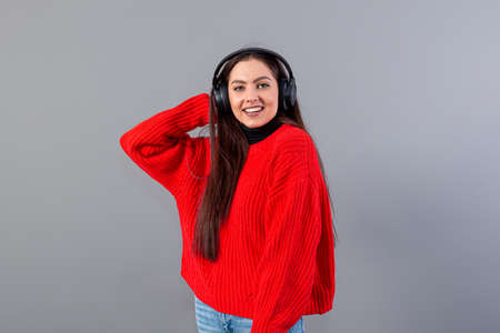 positive teenager brunette in red sweater listens to music on smartphone in headphones, isolated on...