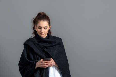 curly-haired emotional young woman in a black cloak using mobile phone, isolated on gray background