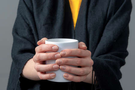 woman in a black cloak is holding white mug with hot drink in both hands, close-up