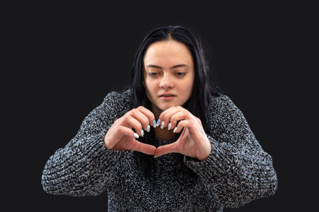 teenager with long dark hair making heart shape hand gesture and looking at camera, isolated on blackの写真素材
