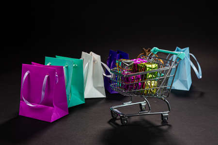 small metal shopping cart full of gifts, colorful bags, isolated on dark, shopping online, winter sale, supermarket, discount promotion and black Friday conceptの写真素材