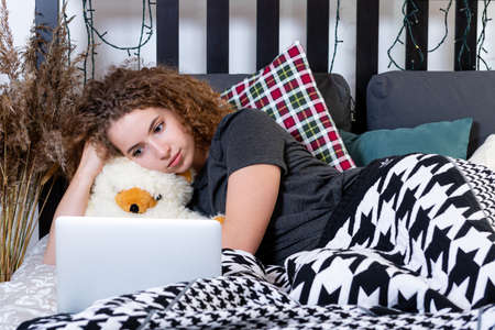 a young curly teen girl lies in bed caught a teddy bear and looks at the laptop screen