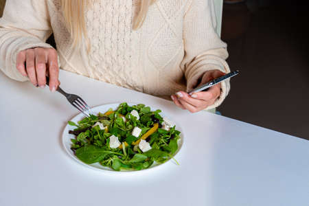 middle aged blonde woman  using smartphone while eating salad in the kitchen, lifestyle concept, close-upの写真素材