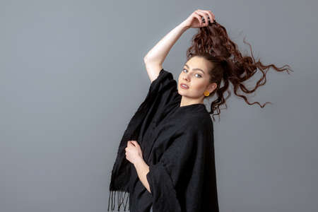 portrait of charming curly-haired teenager woman in a black cloak isolated on a gray background