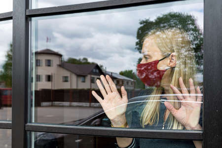 depresive woman in face mask looking out the window, staying home in quarantine, covid-19 lockdown