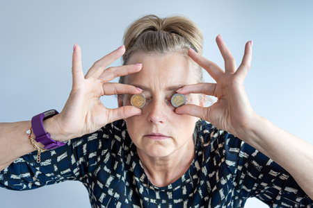 an middle-aged woman holds euro coins in her hands near her eyes, closeup headshot