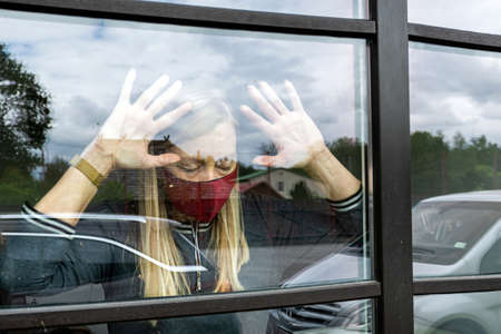 depresive woman in face mask looking out the window, staying home in quarantine.