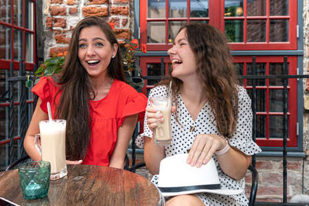 two cheerful pretty young women drinking coffee, talking and laughing in outdoor cafe