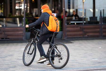 Riga, Latvia - September 3, 2021: a young man in a brightly colored hat and with a backpack on a bicycle in a paved area near a cafeのeditorial素材