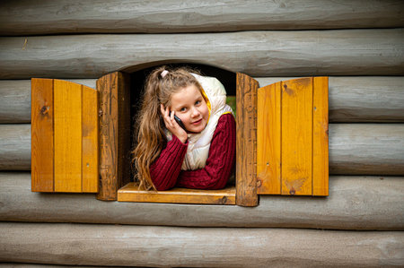 The cheerful little girl looks out the window of a wooden log house and talks on a smartphoneの写真素材