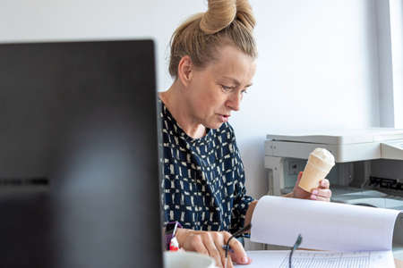 a busy middle-aged office woman eats ice cream at her workplace without interrupting work