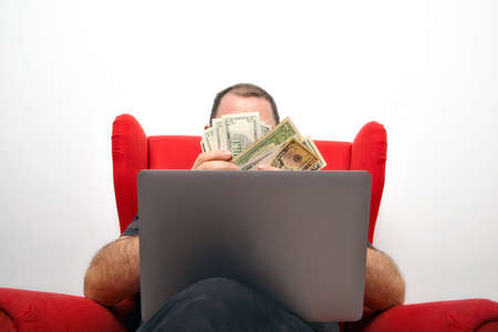 middle-aged man in eyeglasses with a laptop on his lap sitting in a red chair and counting moneyの写真素材